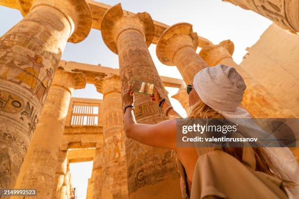female tourist walks between pillars of the great hypostyle hall from karnak temple, and taking pictures with smart phone - templo de amon imagens e fotografias de stock