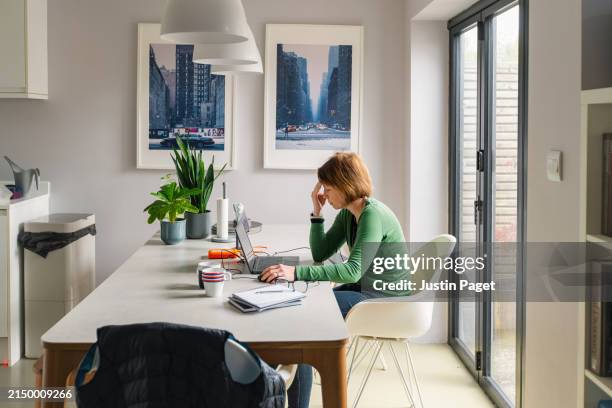 a stressed mature woman works from her dining table at home - espacio de trabajo virtual compartido fotografías e imágenes de stock