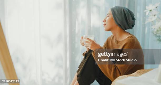 thoughtful young asian woman sick with cancer in a headscarf enjoys a quiet moment with a warm tea, sitting by a sunlit window at home. living with cancer. - oncologia imagens e fotografias de stock