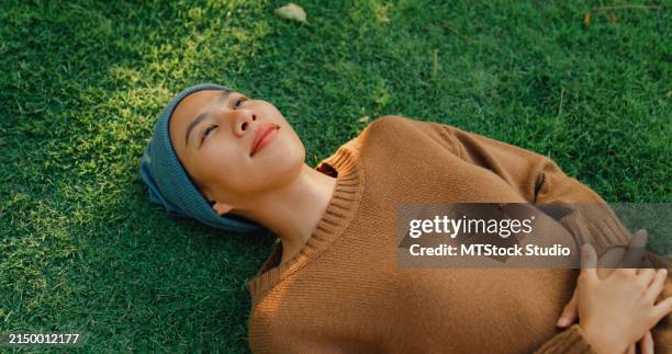 closeup of young asian woman sick with cancer rests on grass, taking a moment to relax and breathe in the peaceful atmosphere of sunny park, showcasing a serene lifestyle. living with cancer. - bildtechnik stock-fotos und bilder