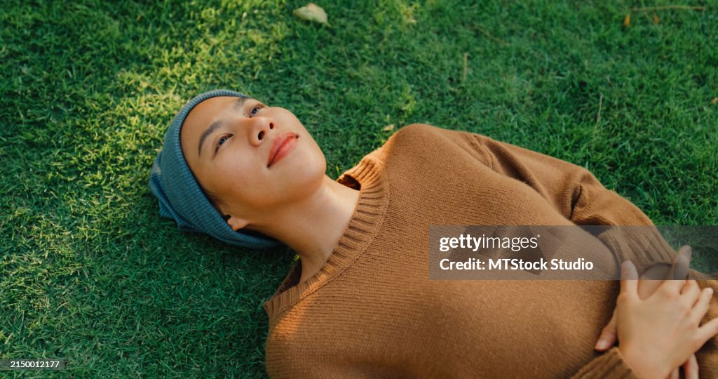 Nahaufnahme einer jungen asiatischen Frau, die an Krebs erkrankt ist, ruht sich auf Gras und nimmt sich einen Moment Zeit, um sich zu entspannen und die friedliche Atmosphäre des sonnigen Parks zu atmen, der einen ruhigen Lebensstil zeigt. Leben mit Krebs