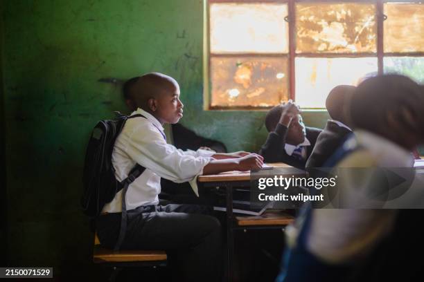 side view male african student sitting in wooden desk listening in a rural classroom - developing countries stock pictures, royalty-free photos & images