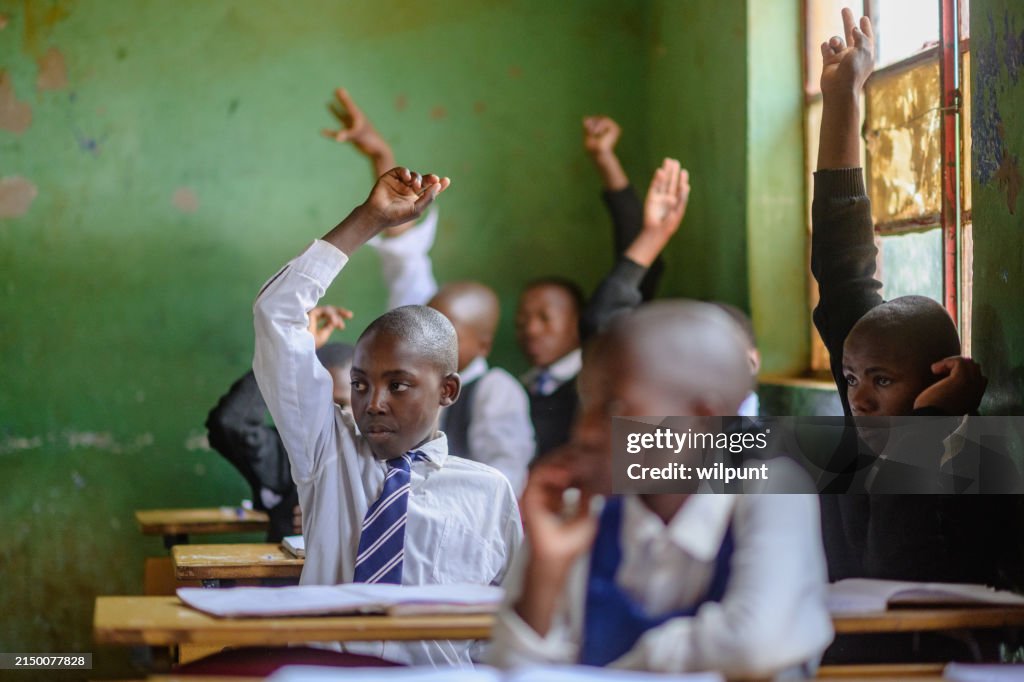Group of African learners wearing school uniform with their hands raised in a classroom