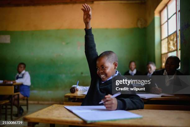 cute schoolboy in uniform sitting in classroom hand raised smiling looking away - developing countries stock pictures, royalty-free photos & images