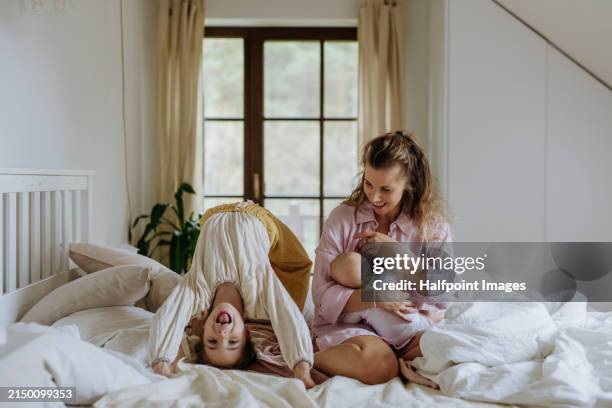 mother breastfeeding baby girl, laughing with older daughter doing gymnastic on bed. perfect mom, perfect moment. - baby good morning stock pictures, royalty-free photos & images