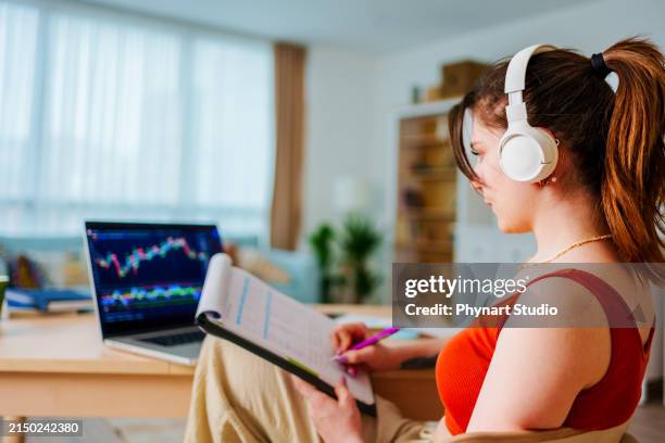 junge geschäftsfrau mit kopfhörern, die bitcoin oder den börsenkurschart an der digitalen börse auf einem laptop-monitor an ihrem schreibtisch zu hause überprüft - crowdsourcing stock-fotos und bilder