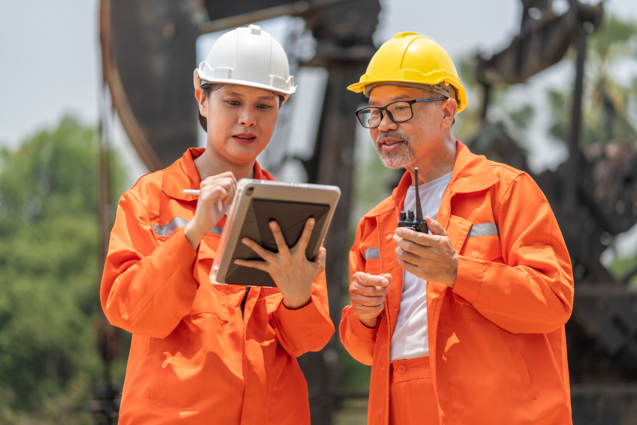 Female engineer wearing white hardhat and orange jumpsuit holding tablet working with a middle age male technician while both standing in front of an oil driller Female engineer wearing white hardhat and orange jumpsuit holding tablet working with a middle age male technician while both standing in front of an oil driller