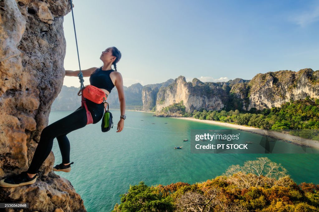 A woman is rock climbing on a mountain with a backdrop of mountains, the sea, a beach and a long-tailed boat.