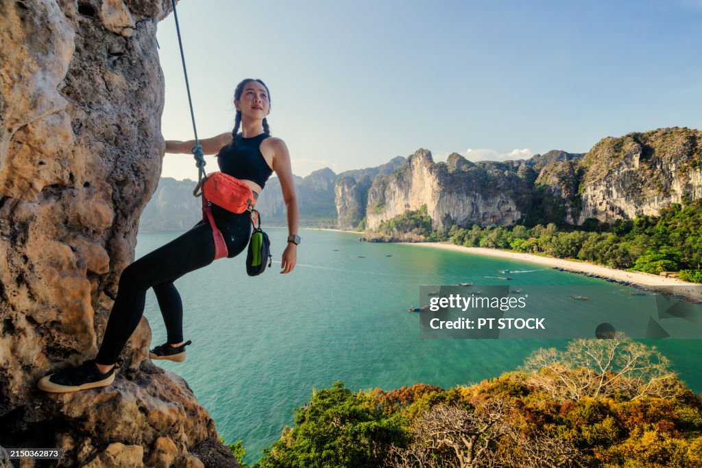 A woman is rock climbing on a mountain with a backdrop of mountains, the sea, a beach and a long-tailed boat.