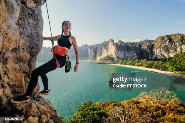 a woman is rock climbing on a mountain with a backdrop of mountains, the sea, a beach and a long-tailed boat. - rock climbing stock pictures, royalty-free photos & images