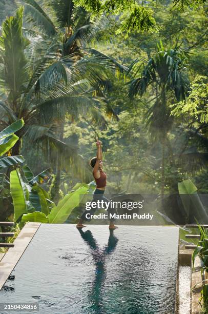 mature fit asian woman practicing yoga on the edge of an infinity pool with early morning mist rising from jungle - bali fotografías e imágenes de stock