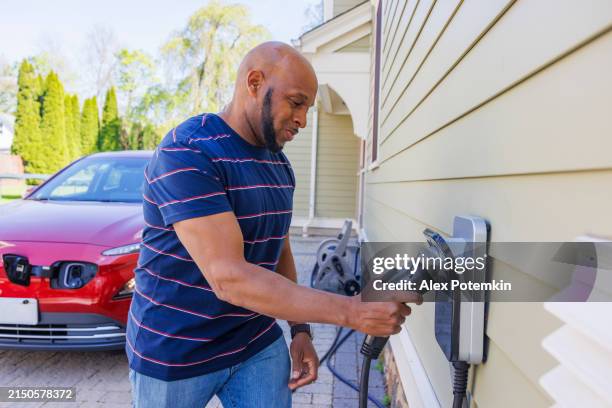 utilisation des énergies renouvelables : un afro-américain heureux est vu en train de brancher un câble de recharge sur un chargeur de véhicule électrique installé dans sa maison de banlieue à madison, dans le new jersey - chargeur de batterie photos et images de collection