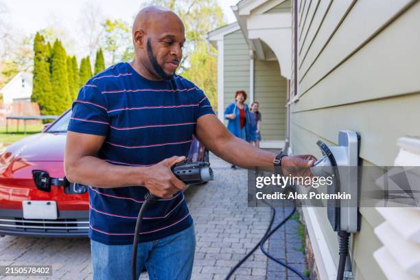 family responsible driving with electric vehicles. african-american father holding ev charger, ready to plug it into red car outside their home in madison, nj - batterijoplader stockfoto's en -beelden