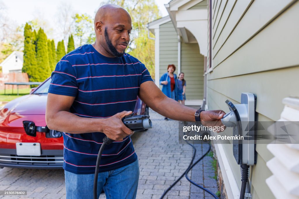 Family responsible driving with electric vehicles. African-American father holding EV charger, ready to plug it into red car outside their home in Madison, NJ
