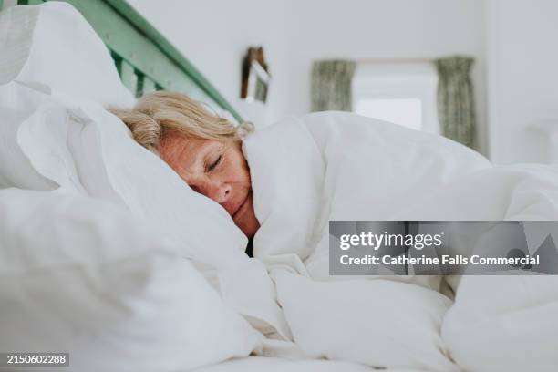 a mature woman snuggles up in a clean, white duvet in a bright and airy bedroom - acolchado fotografías e imágenes de stock