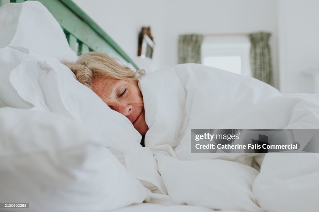 A mature woman snuggles up in a clean, white duvet in a bright and airy bedroom