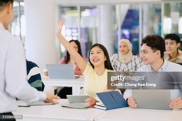 happy student raising arm to answer question while attending class with her university colleagues. - asian students classroom computer stock pictures, royalty-free photos & images