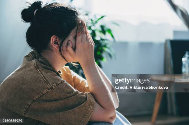 upset teen girl sit on floor sadly look out window worried about teenage problem at school and communication with parent. worried girl tensely suffer about bullying at school, unrequited love with boy - abuso fotografías e imágenes de stock