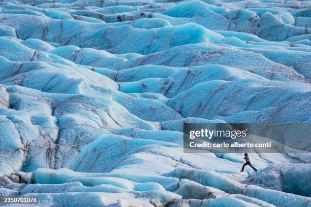 a person has fun and jumps between the particular textures of the ice formed in the svinafellsjokull glacier, affected by the slow melting of the ice due to global warming, iceland, europe - reiseroute golden circle stock-fotos und bilder