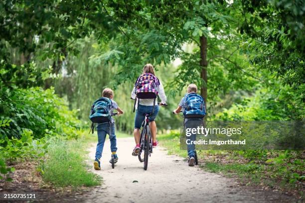 tres niños van a la escuela en bicicleta y patinetes por el parque público - movilidad sostenible fotografías e imágenes de stock