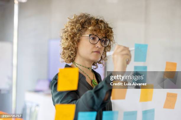 businesswoman writing on adhesive note on glass wall at startup workplace - market research stock pictures, royalty-free photos & images