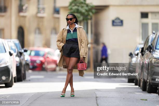 Emilie Joseph wears sunglasses, golden earrings and necklace, a green and blue striped polo shirt, a white shirt, a beige oversized trench coat,...