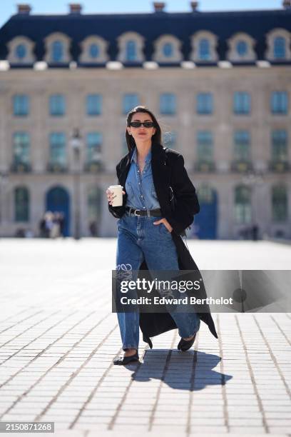Alexandra Pereira wears sunglasses, a black long trench coat , a blue denim shirt, a belt, blue denim pants / jeans with embroidered logo from Loewe,...