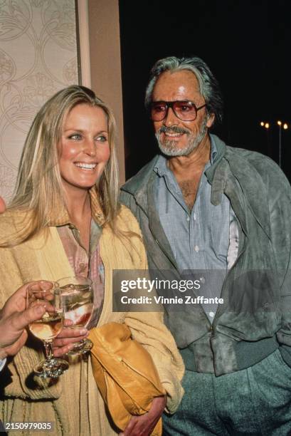 American actress Bo Derek, wearing a yellow coat and carrying a yellow purse, and her husband, American actor and film director John Derek, who wears...
