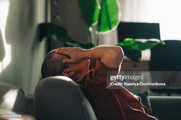 unpleasant pain. sad unhappy handsome man sitting on the sofa and holding his forehead while having headache - disperazione foto e immagini stock