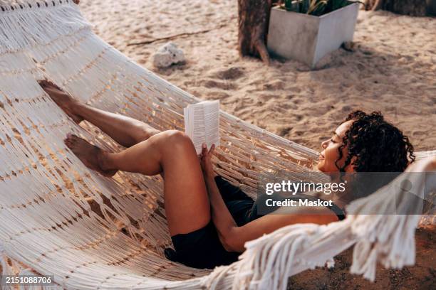 woman reading book lying down in hammock on vacation at beach - hammock stock pictures, royalty-free photos & images
