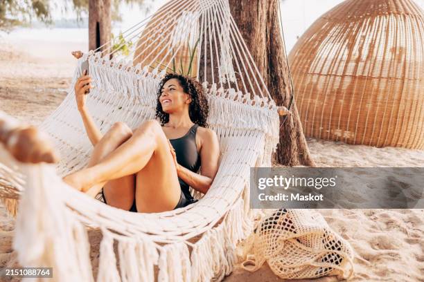 smiling woman doing video on smart phone while lying down in hammock at beach - vacanza al mare foto e immagini stock
