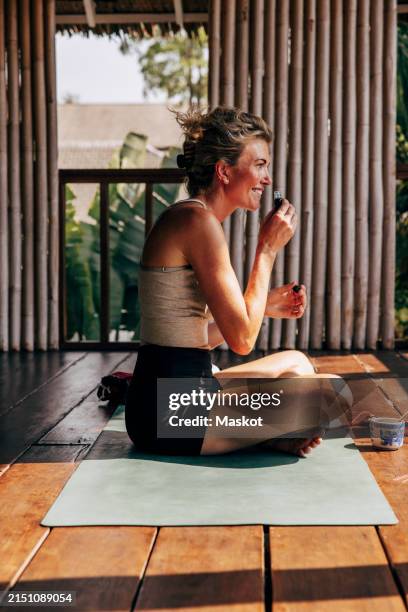 side view of smiling woman smelling oil while sitting on yoga mat at wellness resort - aromaöl stock-fotos und bilder