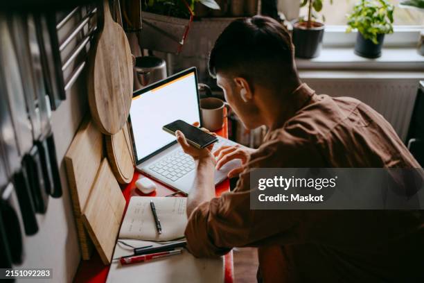 high angle view of male freelancer talking on smart phone while using laptop in kitchen at home - speech recognition stock pictures, royalty-free photos & images