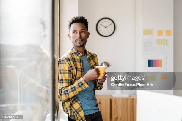 african man drinking coffee looking through the window in the home office - three quarter length stock pictures, royalty-free photos & images