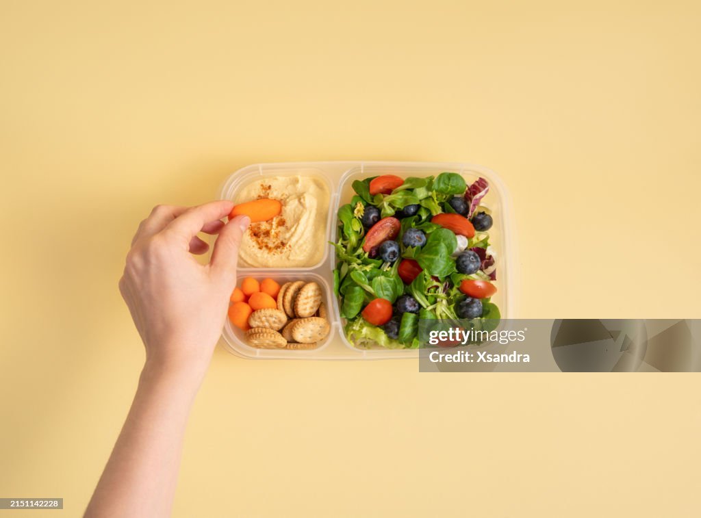 Hand eating baby carrot from a plastic container with hummus and healthy salad