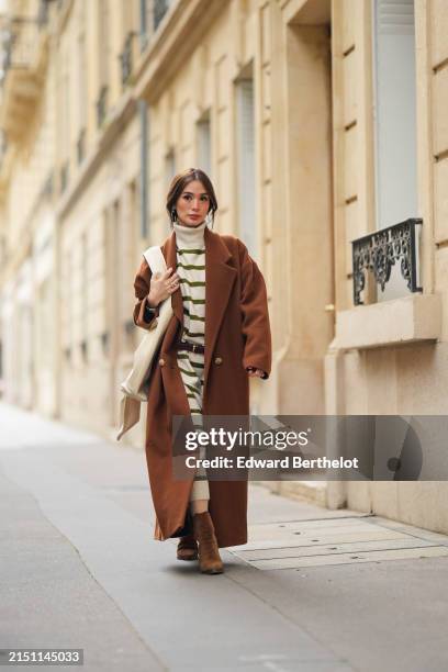 Heart Evangelista wears earrings, a brown long oversize trench coat, a black and white turtleneck striped long dress, a leather belt, a white leather...