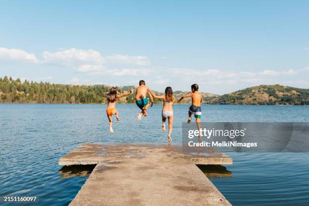 children having fun jumping from a pier in to the lake on a hot summer day - group-of-friends-jumping-off-dock-into-lake stock pictures, royalty-free photos & images