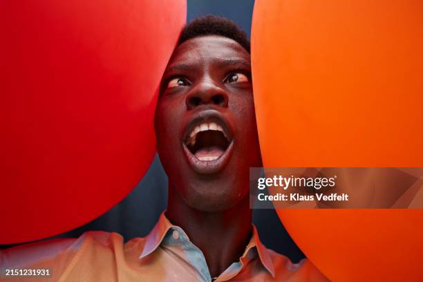 young man making face amidst red and orange balloons - tordu photos et images de collection