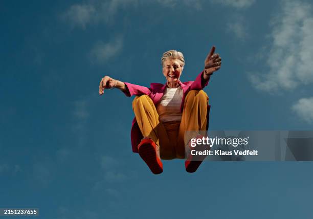 happy senior woman crouching mid-air against blue sky - femmes seniors photos et images de collection
