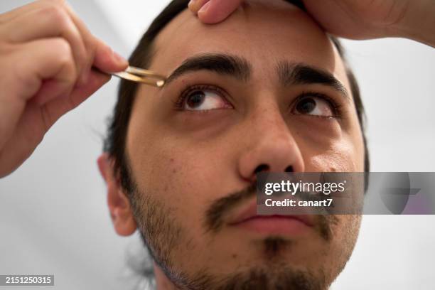 young man is plucking his eyebrows with tweezers - tweezing eyebrows stock pictures, royalty-free photos & images