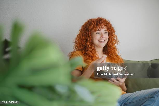 curly haired young woman sitting on the couch and eating popcorn while watching her favorite streaming service on her tv. - streaming service stock pictures, royalty-free photos & images