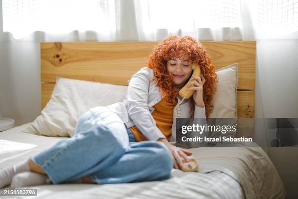 young woman with curly hair talking on the landline phone with her friends lying on her bed. - telefone fixo imagens e fotografias de stock