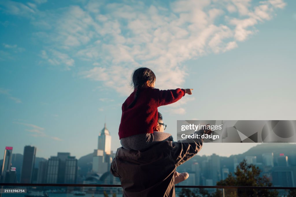 Loving Asian father carrying little daughter on shoulders, pointing far away while looking over city skyline against the promenade of Victoria harbour on a sunny day. Enjoying father-daughter bonding time together. Family love and togetherness lifestyle
