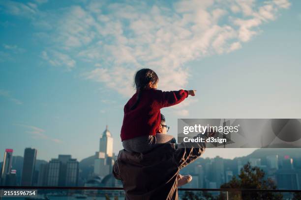 loving asian father carrying little daughter on shoulders, pointing far away while looking over city skyline against the promenade of victoria harbour on a sunny day. enjoying father-daughter bonding time together. family love and togetherness lifestyle - anticipation stock pictures, royalty-free photos & images