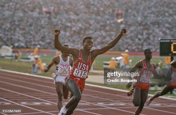 American athlete Carl Lewis, wearing a red Kappa singlet, raises his arms in triumph, competes at the 1984 Summer Olympics, at the Los Angeles...