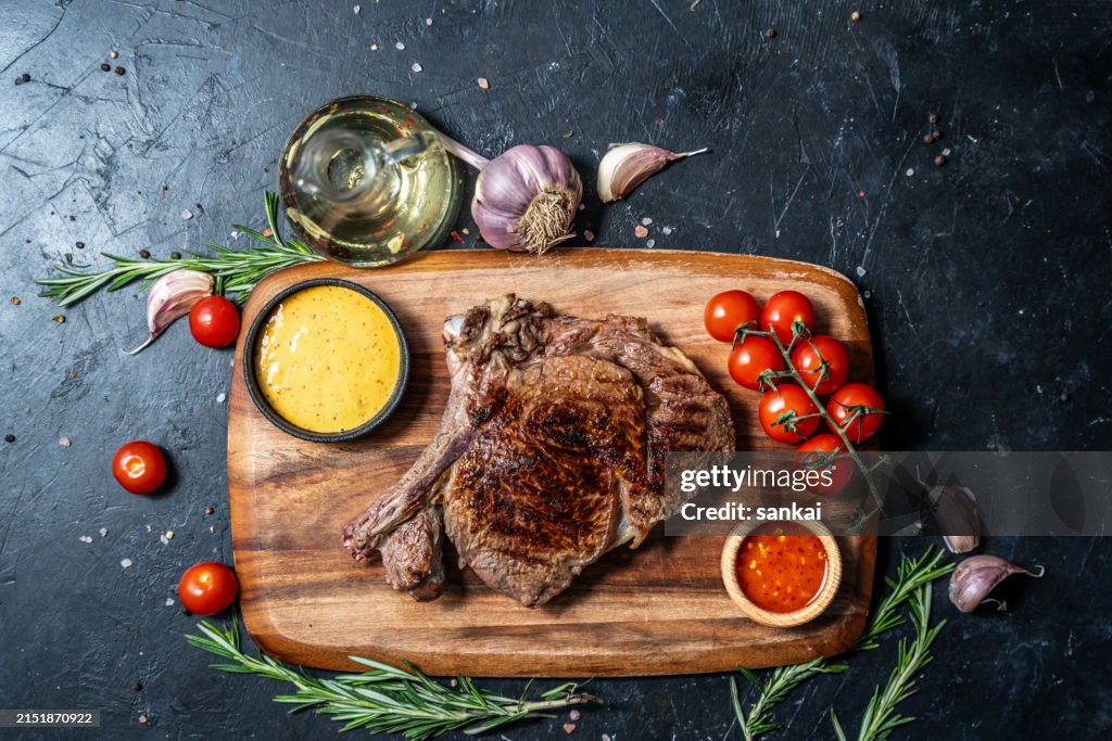 Sabroso filete de ternera a la parrilla con hueso sobre tabla de madera con salsa de chile dulce y salsa de mostaza, decorado con tomates cherry, especias y hierbas.