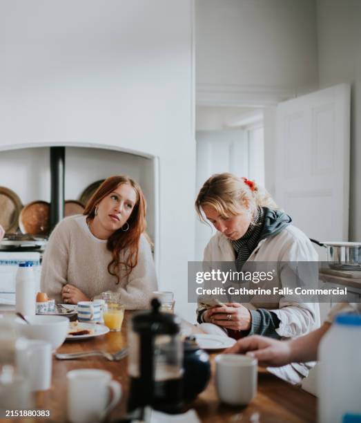 a young woman is distracted by her smart phone at a kitchen table - sibling stock pictures, royalty-free photos & images