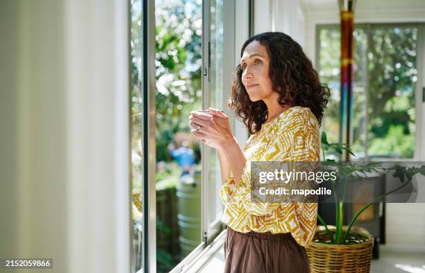 donna matura che beve caffè mentre guarda fuori attraverso una finestra di casa - guardare fuori dalla finestra foto e immagini stock