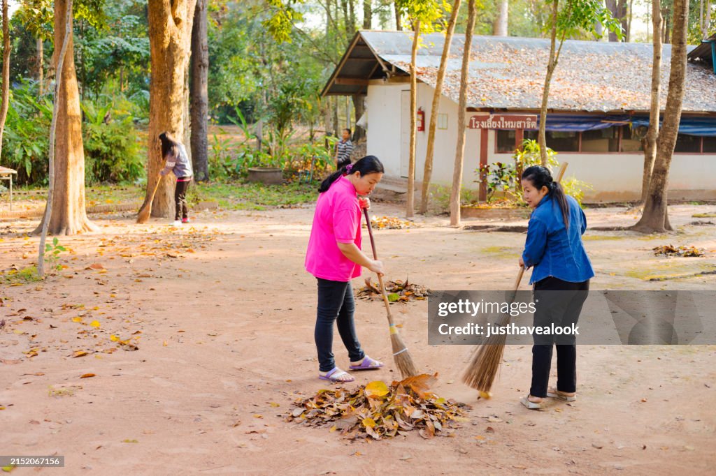 Two adult Thai woman are cleaning remote location road with broom