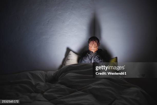 depressed man covering face with hands sitting on bed in dark room - un solo hombre fotografías e imágenes de stock